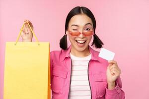 Lycklig ung asiatisk kvinna som visar kreditera kort för handla, innehav väska, uppköp på försäljning, gående till de affär, Lagra, stående över rosa bakgrund foto