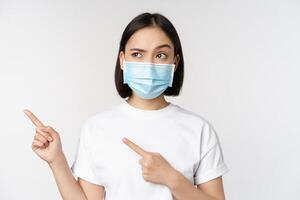 bild av asiatisk kvinna i medicinsk mask från covid, ser förvirrad vänster, pekande på logotyp, som visar annons, stående över vit bakgrund foto