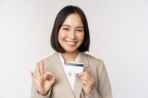 leende kontor kontorist, asiatisk företags- kvinna som visar kreditera kort och Okej tecken, rekommenderar Bank, stående över vit bakgrund i beige kostym foto