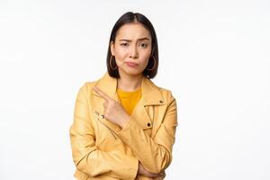 porträtt av eleganta asiatisk flicka ser med tvivel, skeptisk ansikte, pekande finger vänster på logotyp eller annons, stående mot vit bakgrund foto