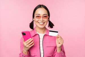 uppkopplad handla. leende asiatisk flicka shoppare, innehav smartphone och kreditera kort, betalande i mobil app, stående över rosa bakgrund foto