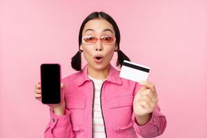 asiatisk flicka visar mobil telefon skärm och kreditera kort, reagerar överraskad på kamera, gasning imponerad, stående över rosa bakgrund, uppkopplad handla begrepp foto