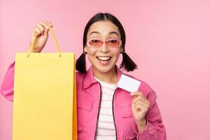 Lycklig ung asiatisk kvinna som visar kreditera kort för handla, innehav väska, uppköp på försäljning, gående till de affär, Lagra, stående över rosa bakgrund foto