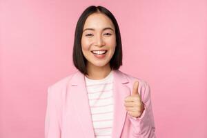 porträtt av asiatisk affärskvinna leende nöjd, som visar tummen upp, beröm, tycka om och godkänna, stående i kostym över rosa bakgrund foto