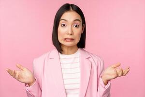 porträtt av förvirrad asiatisk affärskvinna rycker på axlarna axlar, ser aningslös och förbryllad, gör inte känna till, kan inte säga, stående över rosa bakgrund i kontor kostym foto