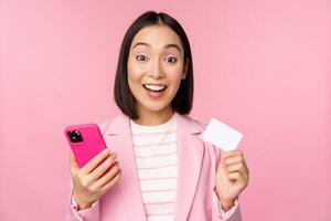 bild av leende Lycklig asiatisk affärskvinna som visar kreditera kort, betalande uppkopplad på smartphone Ansökan, beställa med mobil telefon, stående mot rosa bakgrund foto