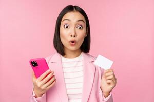 förvånad affärskvinna, asiatisk flicka i kostym som visar kreditera kort och mobil telefon, beställa uppkopplad, handla med smartphone, stående över rosa bakgrund. reklam begrepp foto