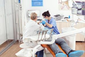 sjukhus tandläkare team rengöring tänder av sjuk man framställning för stomatologi kirurgi under stomatologisk inspektion. patient Sammanträde på dental stol väntar för ortodontisk inspektion foto