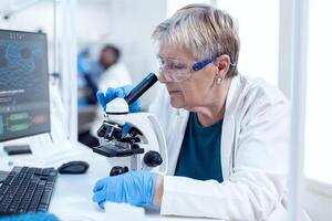 senior kemist forskare håller på med genetisk undersökning använder sig av mikroskop. äldre forskare bärande ut vetenskaplig forskning i en steril labb använder sig av en modern teknologi. foto