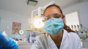 patient pOV besöker dental klinik för kirurgi behandla påverkade massa. läkare och sjuksköterska arbetssätt tillsammans i modern ortodontisk kontor, belysning de lampa och granskning person bär skydd mask. foto