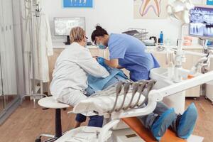 sjuk man Sammanträde på dental stol under medicinsk undersökning medan senior tandläkare kvinna håller på med oral kirurgi i tandvård kontor. sjukhus team granskning patient tandvärk framställning tand behandling foto