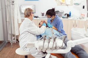 senior kvinna tandläkare analyserar tänder granskning sjuk patient medan Sammanträde på ortodontisk stol. läkare stomatolog och sjuksköterska rengöring mun under dental undersökning i stomatologi klinik foto