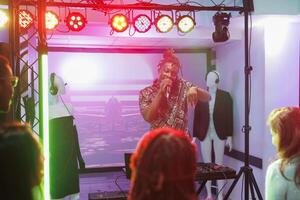afrikansk amerikan dj sång och spelar på skede under fest i nattklubb. ung man musiker utför och blandning musik på diskotek i klubb med spotlights på natt foto