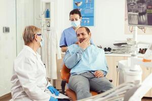 sjuk patient klagar handla om tandvärk medan talande med tandläkare innan ingripande. senior läkare tala till man Sammanträde på stomatologisk stol medan sjuksköterska stomatologi sjukhus kontor foto