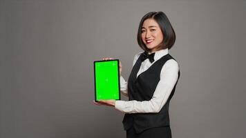 kvinna concierge presenter grönskärm visa på läsplatta, som visar Chromakey mall på skärm. receptionist med rosett och enhetlig innehav enhet med tom copy layout. kamera a. foto