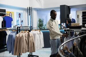 afrikansk amerikan man bläddring kläder på kuggstång medan handla i eleganta boutique. kund sökande för modern modern formell ha på sig på galge i detaljhandeln Centrum utställningslokal foto