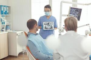 ortodontist sjuksköterska innehav digital läsplatta med tand radiografi på skärm förklara till sjuk man stomatologi behandling till förhindra tandvärk. patient Sammanträde på dental stol i modern tandvård kontor foto