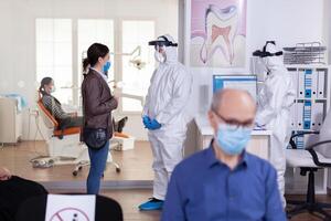 tandläkare läkare klädd i ppe kostym diskuterar med patient i stomatologi reception korridor under utnämning , i tid av global pandemi med coronavirus hälsa kris. foto