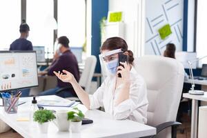 företag kvinna under global pandemi med covid 19 har en företag konversation använder sig av smartphone bär ansikte mask. multietnisk medarbetare arbetssätt respekterar social distans i finansiell företag. foto