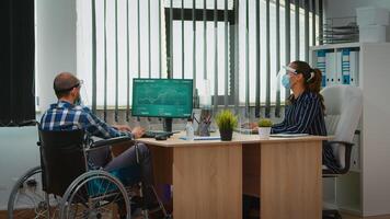 chef med handikapp comming med rullstol på arbetsplats med skydd mask arbetssätt i ny vanligt företag kontor. immobiliserad frilansare i finansiell företag respekterar social distans. foto