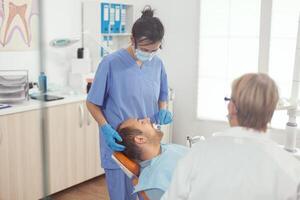 medicinsk assistent kvinna håller på med dental behandling till sjuk man framställning stomatologi verktyg för dental kirurgi. man lokaliserings på tandvård stol i sjukhus ortodontisk kontor under medicinsk procedur foto