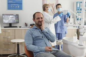 porträtt av man patient med tänder smärta Sammanträde på dental stol under stomatologisk undersökning väntar för medicinsk expertis. i bakgrund specialist team diskuterar tandvård behandling foto