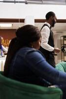 afrikansk amerikan hotell personal och gäst i lobby. kvinna resande väntar för checka in på reception område, vänlig leende bellboy portion gäster med bagage. gästfrihet och turism industri foto