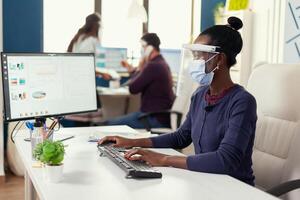 afrikansk kvinna skriver på dator på arbetsplats bär ansikte mask som säkerhet försiktighetsåtgärd mot covid19. multietnisk team i ny vanligt företag finansiell kontor kontroll rapporter, analyserar data ser på skrivbordet. foto