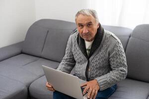 äldre man Sammanträde på soffa, leende på dator skärm på Hem foto