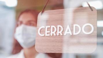 ung ledsen kvinna med ansiktsmask ändra öppen till stängd skylt på spanska på fönstret för corona-virus foto