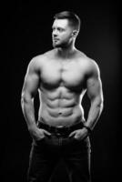 muskulös kille med naken torso Framställ med händer i fickor. studio Foto. porträtt av en stilig man i jeans på mörk bakgrund. svart och vit Foto. närbild. foto