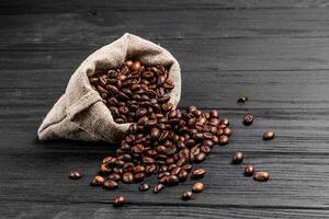 väska av kaffe bönor och några spridd frön på de trä- bakgrund. rostad kaffe korn spill ut av säck på mörk yta. foto