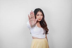 allvarlig asiatisk kvinna bär tillfällig ha på sig med hand gest utgör avslag eller förbud, isolerat förbi vit bakgrund foto