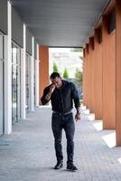 stilig sportigt kille gående genom de byggnad. afrikansk man i svart skjorta och jeans sätta på hans glasögon medan gående. foto