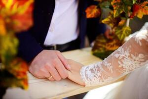 brudgummen hand med gyllene ringa innehar brudens hand på de trä- yta. närbild händer av romantisk par under bröllop ceremoni över de höst löv bakgrund. foto
