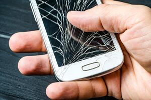 mannens hand innehav mobil telefon med bruten skärm över de mörk bakgrund. smart telefon med glas bruten Rör skärm i mannens hand. närbild foto