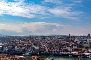 antenn se av en kust stad med historisk byggnader, en floden, och båtar under en blå himmel med moln i whitby, england. foto