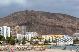 kust stadsbild med vid stranden, höghus byggnader, och en berg bakgrund under molnig himmel i los cristianos, teneriffa. foto