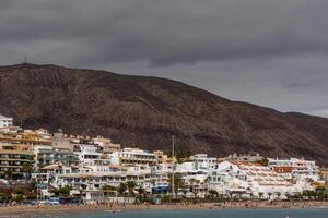 kust stad med vit byggnader inbäddat på de fot av en berg under molnig himmel i los cristianos, teneriffa. foto