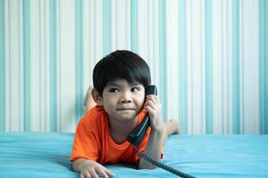 en liten asiatisk pojke lögner på de säng talande på de telefon. foto