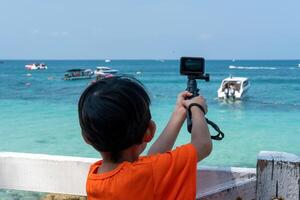 en liten asiatisk pojke är filmning ett uppkopplad video utomhus medan gående till de strand. foto