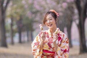 japansk kvinna i traditionell kimono klänning innehav ljuv hanami dango efterrätt medan gående i de parkera på körsbär blomma träd under vår sakura festival foto