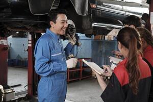 asiatisk manlig professionell bil- ingenjör handledare beskriver bil hjul och suspension reparera arbete med mekaniker arbetstagare staber team i fixera service garage, specialist yrken i bil industri. foto