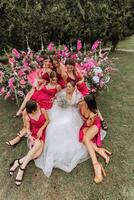 en brunett brud och henne brudtärnor i klänningar av de samma Färg sitta med de brud och glädjas med blommor i deras händer nära de högtidlig båge. bröllop i natur. foto