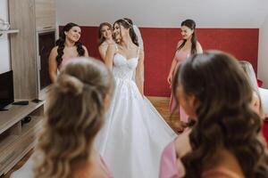 bröllop morgon. brudtärnor hjälp sätta på de vit bröllop klänning. en ung kvinna är framställning till träffa henne brudgum och har roligt med henne vänner foto