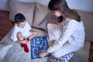 mor med Tonårs dotter och liten son i pyjamas spelar styrelse spel på de golv foto