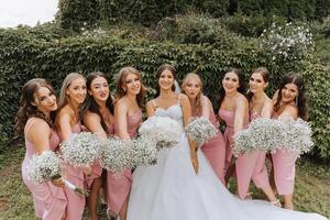 en brunett brud och henne brudtärnor i matchande rosa klänningar är stående med buketter av blommor i deras händer. flickor i identisk klänningar är framställning ut på en bröllop. bröllop i natur. foto