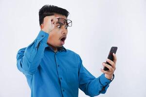 porträtt av överraskad och chockade stilig ung asiatisk affärsman med glasögon i bär blå skjorta använder sig av smartphone och tar av glasögon isolerat på vit bakgrund foto