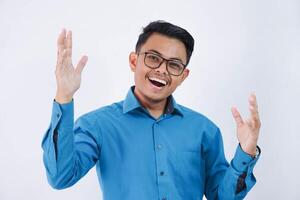 glad eller Lycklig ung stilig asiatisk glasögon man ser kamera med Uppfostrad händer och bär en blå skjorta isolerat på vit bakgrund foto
