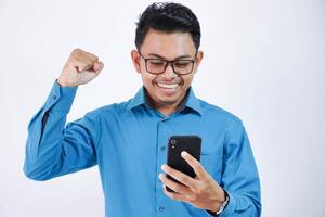 upphetsad ung asiatisk anställd man med glasögon innehav telefon med knöt nävar bär blå skjorta isolerat på vit bakgrund foto